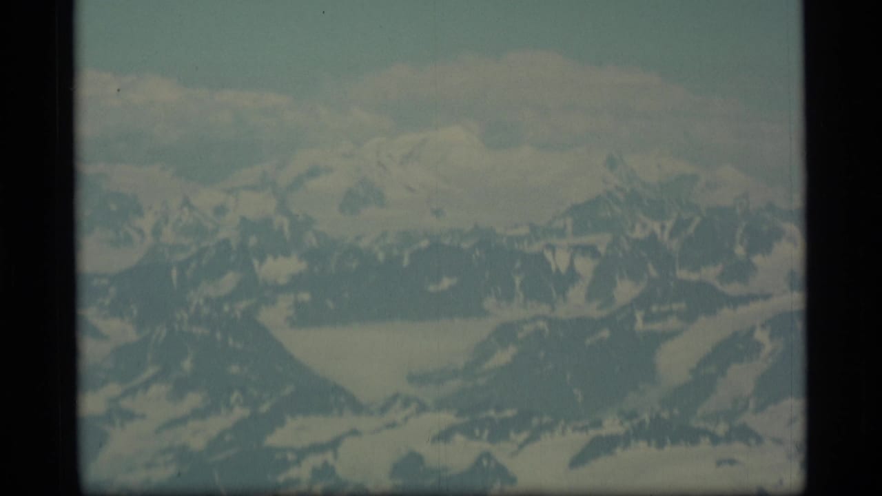 1977 Alaska aerial home-movie frame showing distant snowy peaks, glacier forms, and a pale sky with soft focus and faded color.