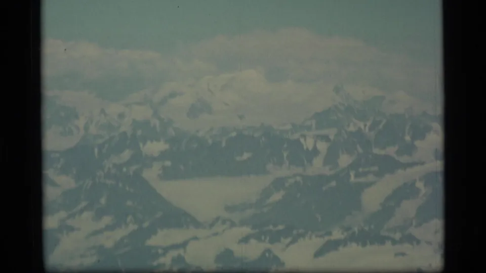 1977 Alaska aerial home-movie frame showing distant snowy peaks, glacier forms, and a pale sky with soft focus and faded color.