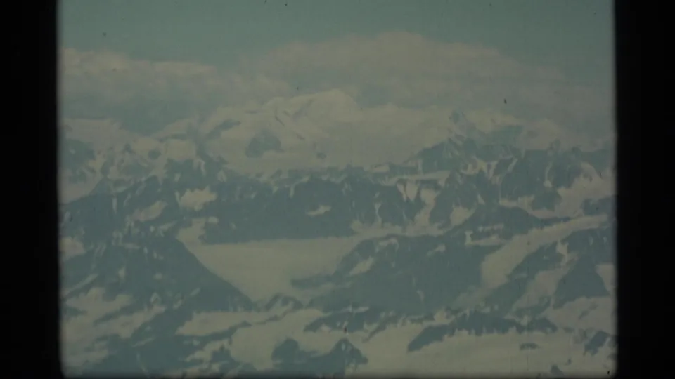 1977 Alaska 8mm aerial frame with broad snow-covered peaks, glacier valleys, muted sky, and visible home-movie film texture.