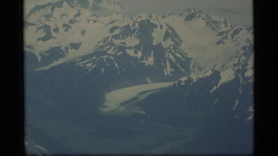 1977 Alaska aerial home-movie frame showing rugged snowy ridges, a glacier tongue, dark lower slopes, and faded 8mm color.