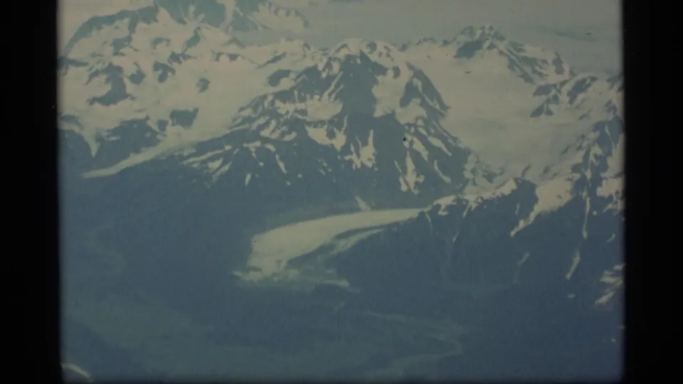 1977 Alaska 8mm aerial frame of snow-streaked mountain ridges, glacier ice, and dark valley terrain under a pale sky.