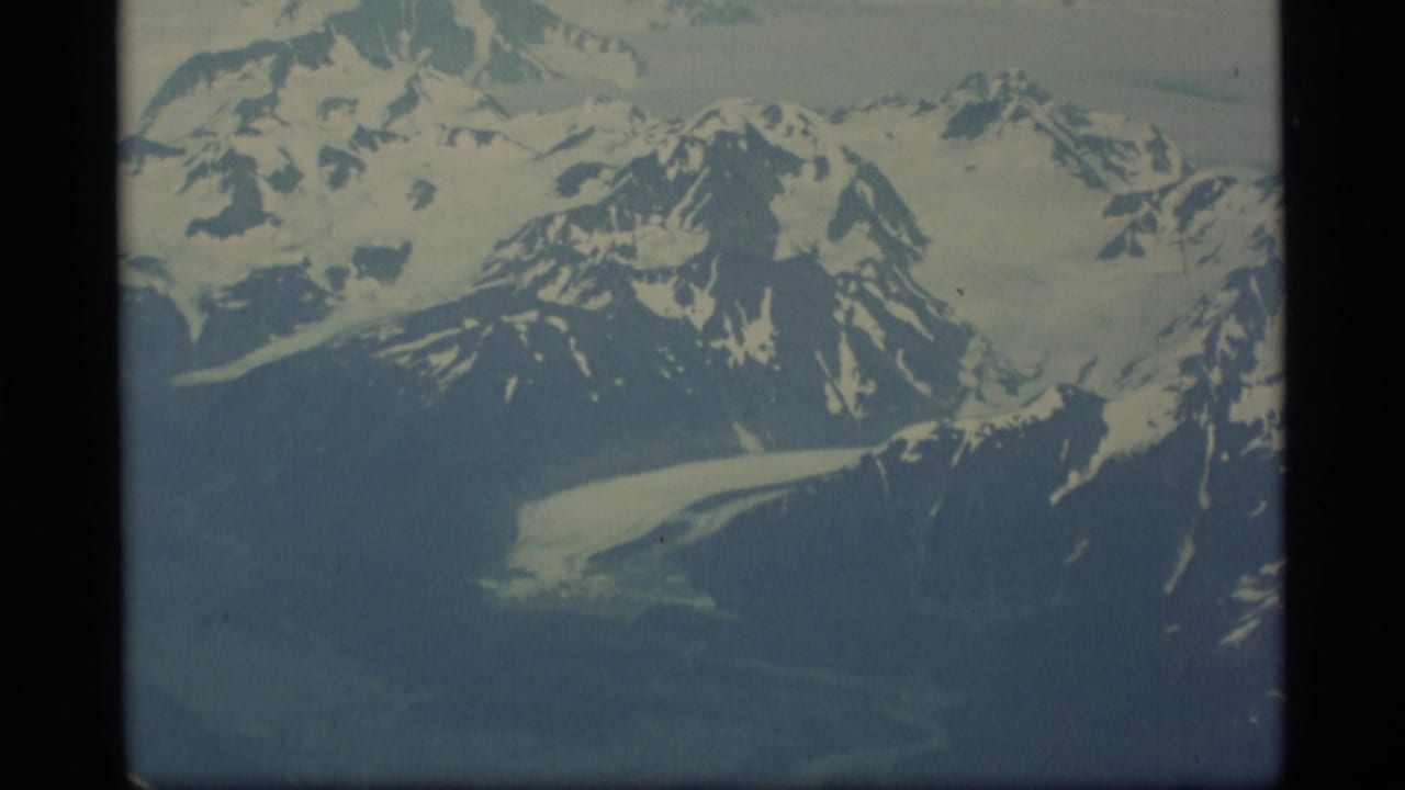 1977 Alaska 8mm frame showing an aerial view of snow-covered mountain ridges and dark rock faces under a hazy sky.