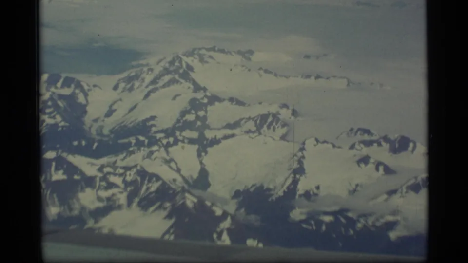 1977 Alaska 8mm frame with snowy mountain masses and glacier-like ice fields seen from an aircraft perspective.