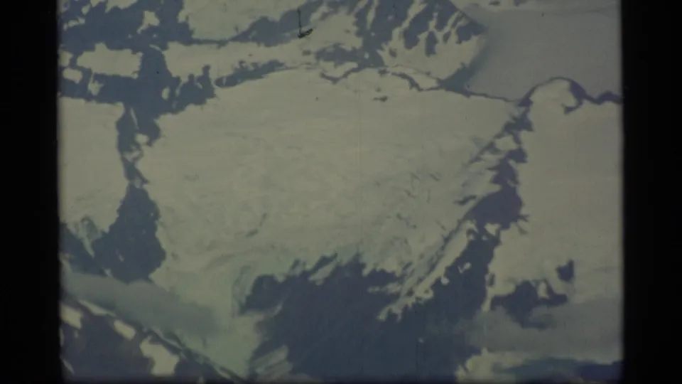 1977 Alaska 8mm frame of a glacier-like ice field below rugged snowy ridges, filmed from an aerial viewpoint.