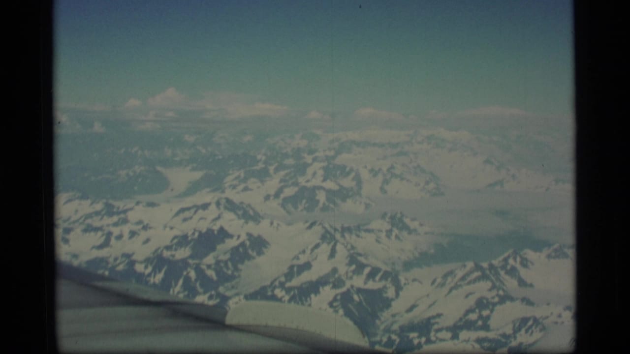 1977 Alaska 8mm home-movie frame showing snowy mountains and glacier-like terrain from an airplane window, with the wing edge visible below.