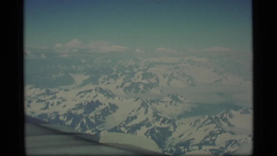 1977 Alaska 8mm home-movie frame showing snowy mountains and glacier-like terrain from an airplane window, with the wing edge visible below.