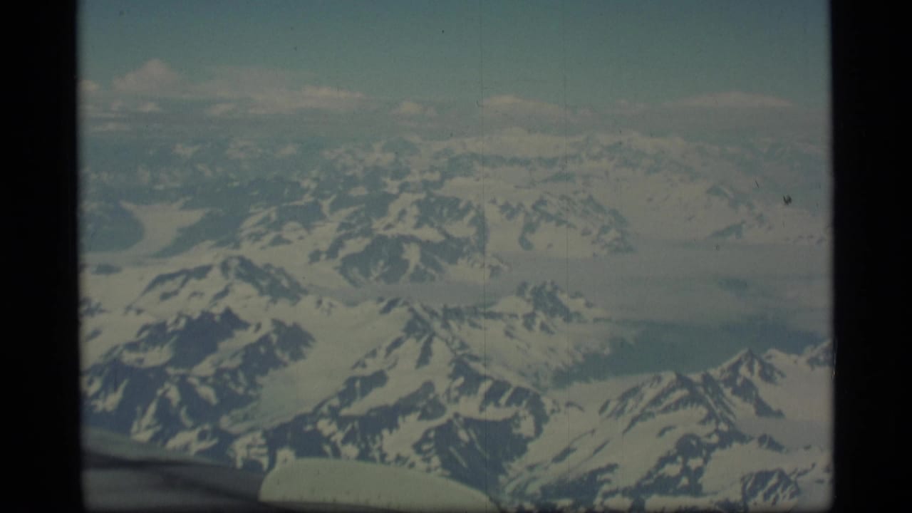 Aerial mountain landscape with snowfields, ridges, pale sky, and a wing edge below.