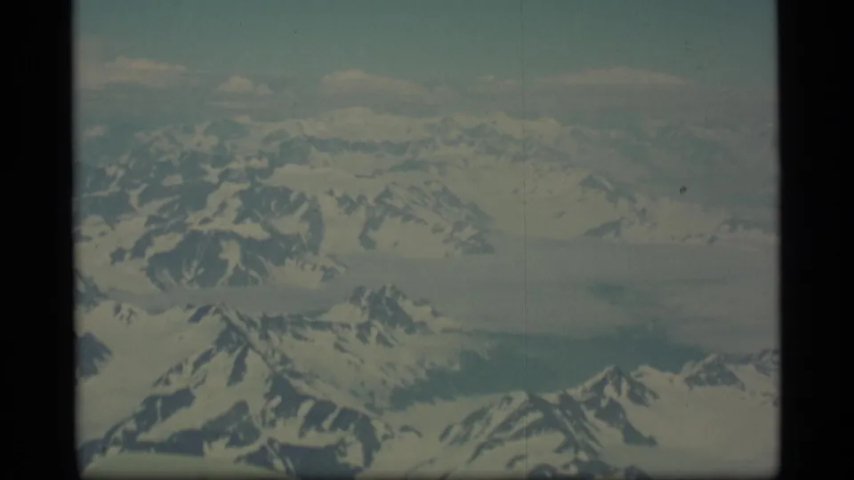 1977 Alaska 8mm frame with a broad aerial view of snow-covered peaks, dark ridges, and glacier-like expanses from a plane.