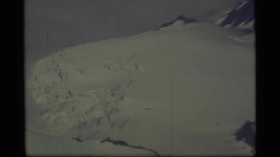 1977 Alaska home-movie frame showing a closer airplane-window view of a large snow-covered slope or glacier surface.