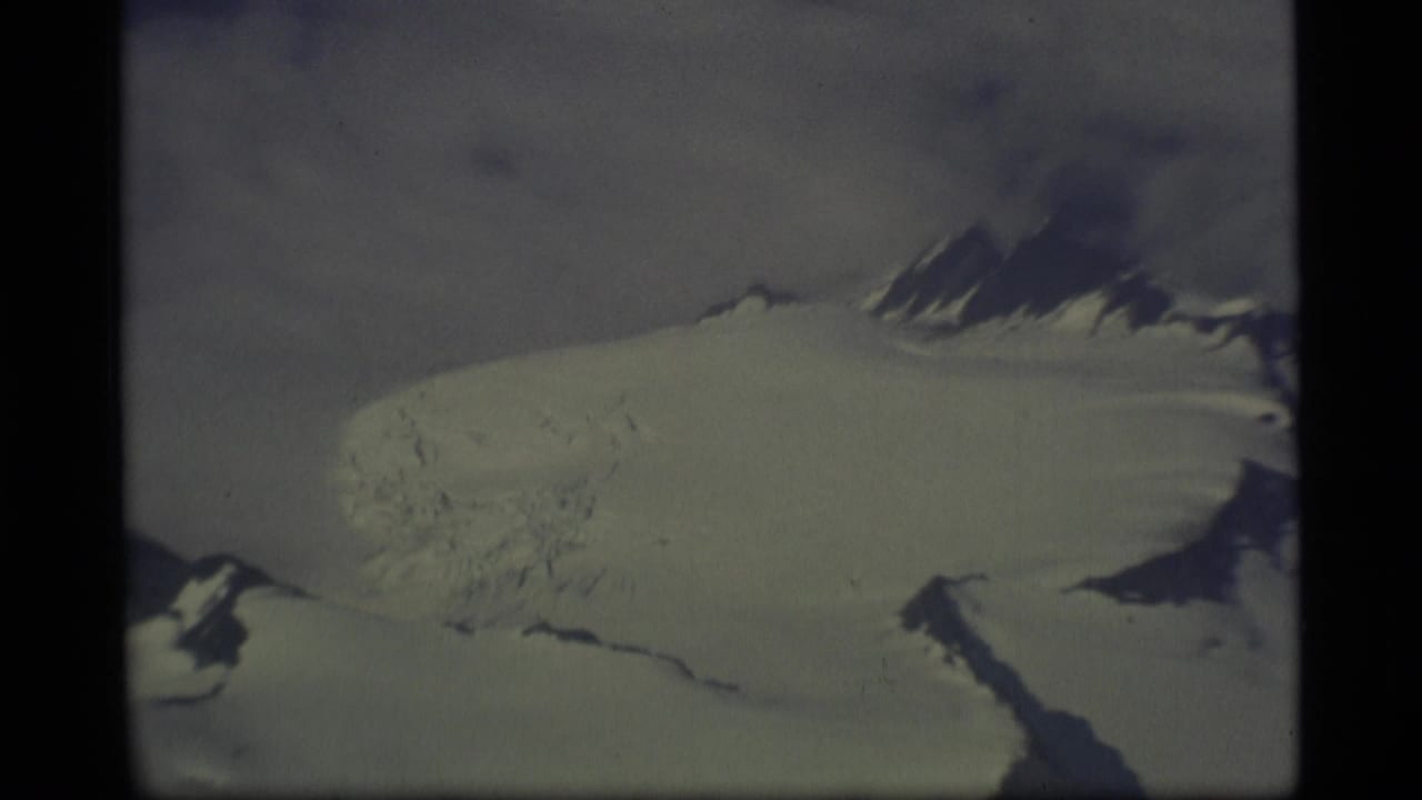 Snow-covered glacier and mountain peaks seen through a dark aircraft window frame.