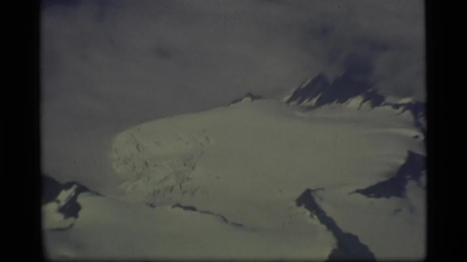 1977 Alaska aerial home-movie view of a broad snowy glacier and dark mountain ridges through an aircraft window.