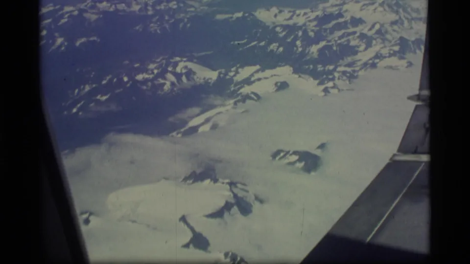Vintage Alaska aerial footage shows the airplane wing crossing a view of snow-covered mountains and glacier terrain.