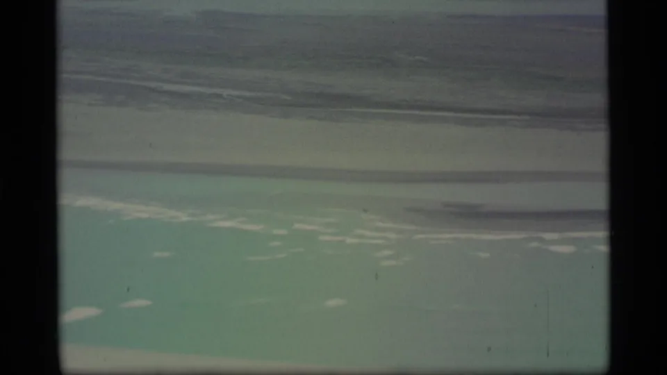 Aerial 8mm view over pale turquoise water or ice near Alaska, with dark cloud or land bands across the upper frame.