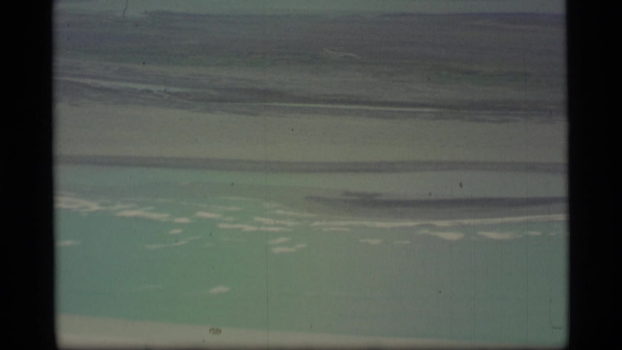 Airplane-window view of pale turquoise water and distant shoreline through a film frame.