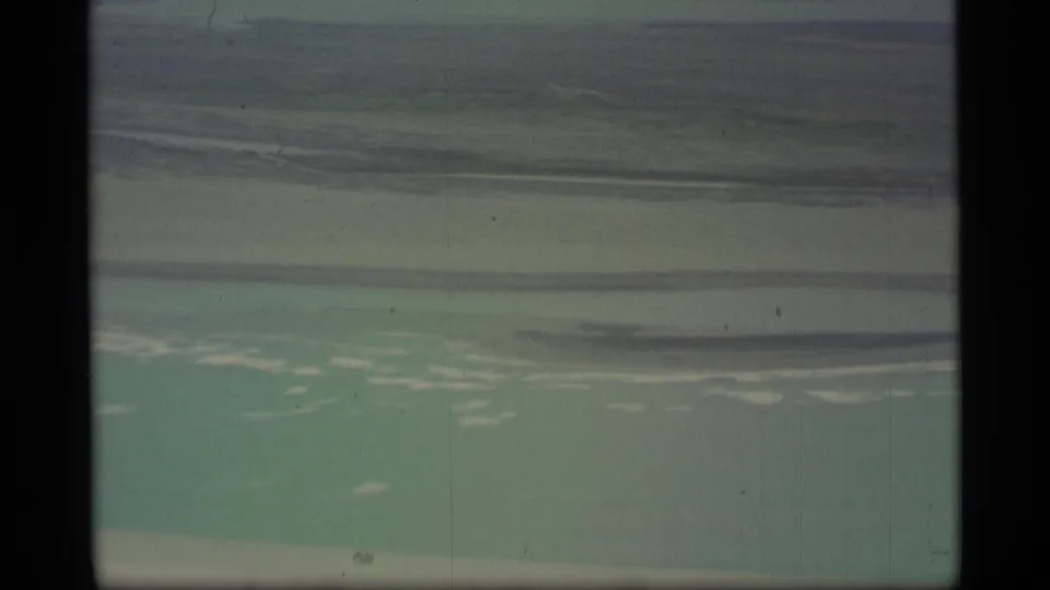 1977 Alaska 8mm frame showing turquoise water, low shoreline, overcast sky, and dark film borders from an airplane-window view.