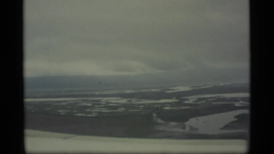 1977 Alaska aerial frame with braided water channels, dark wetland areas, overcast sky, and the aircraft wing edge along the lower frame.