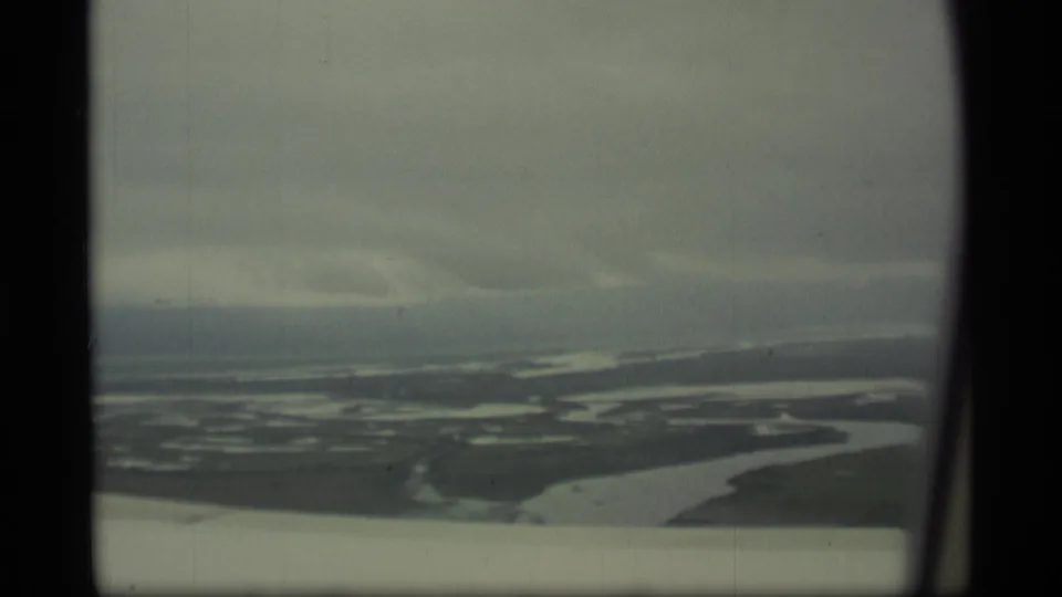 1977 Alaska 8mm aerial view of wetlands, branching water, low terrain, cloudy sky, and a pale aircraft wing edge in the foreground.