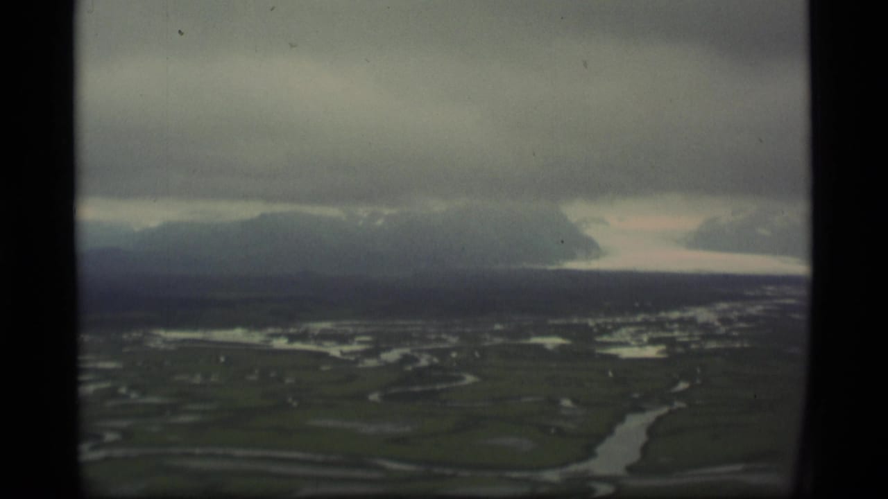 1977 Alaska 8mm aerial view showing green tundra, branching waterways, distant mountains, and heavy clouds through a soft film frame.