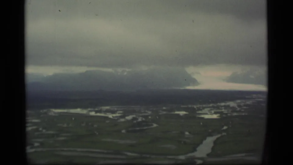 1977 Alaska 8mm aerial view showing green tundra, branching waterways, distant mountains, and heavy clouds through a soft film frame.