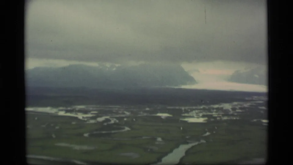 1977 Alaska aerial home-movie frame with winding water channels across tundra and misty mountain ridges below a heavy sky.