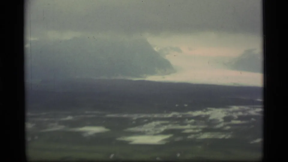 Soft 8mm frame from Alaska in 1977 showing reflective tundra flats, a dark mountain mass, haze, and pale sky from an aircraft viewpoint.