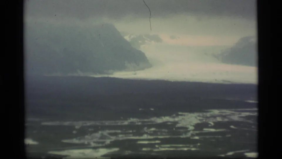 1977 Alaska 8mm aerial frame looking across wet tundra and water patches toward a large cloud-obscured mountain ridge.