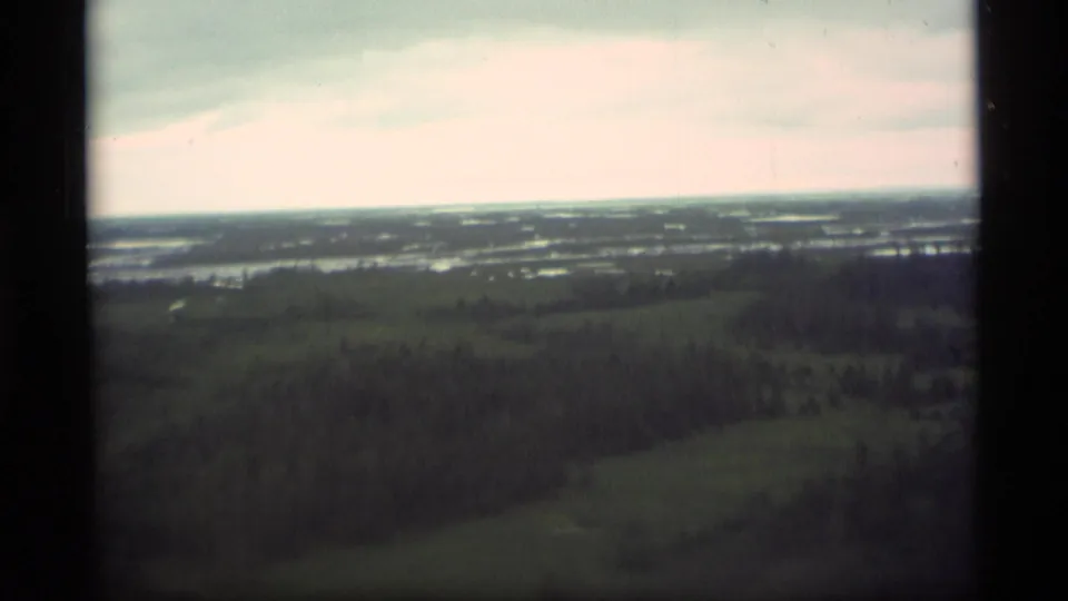1977 Alaska home-movie aerial frame looking across wooded terrain, scattered water, and a cloudy distant horizon.