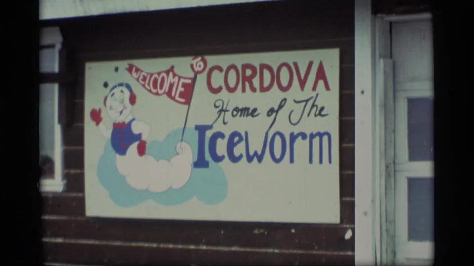 1977 Alaska frame showing a painted Cordova welcome sign with an iceworm mascot mounted on a building exterior.