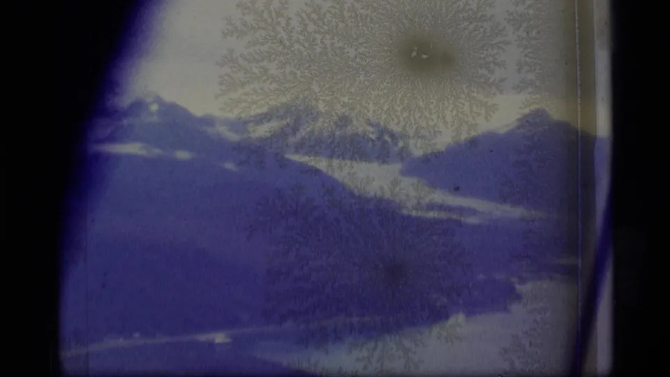 1977 Alaska 8mm frame with a dark lake or valley beneath snowy mountains, partly obscured by blue fading and frost-like texture.