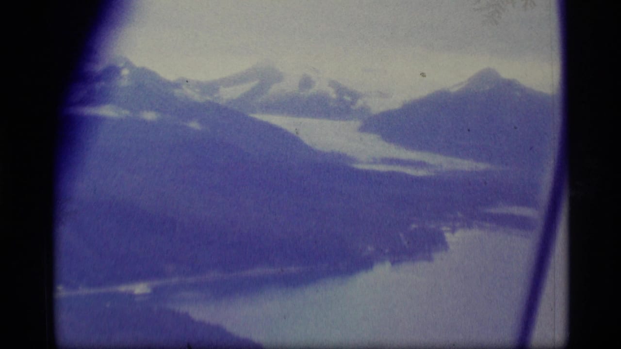 Aerial landscape with snow-capped peaks, dark ridges, and pale water below.