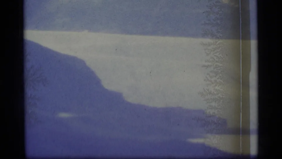 1977 8mm Alaska aerial still showing pale glacial water, dark shoreline shapes, and frost-like marks on the film image.