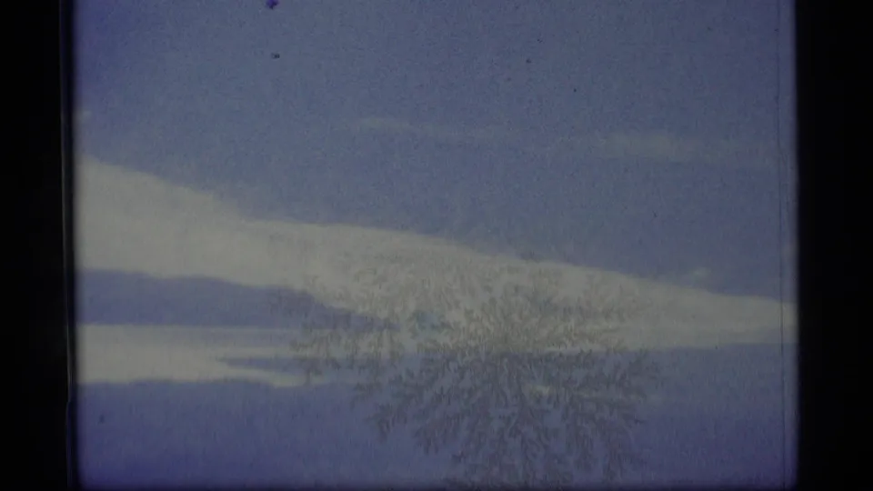 1977 Alaska home-movie frame of a distant snowy ridge and lake, tinted blue and crossed by frost-like branches.