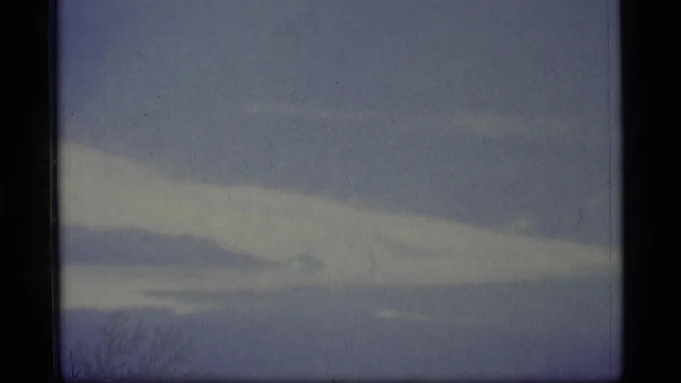 Soft 1977 Alaska 8mm landscape frame with cloud bands, faint mountain forms, water, and pale blue-white exposure.