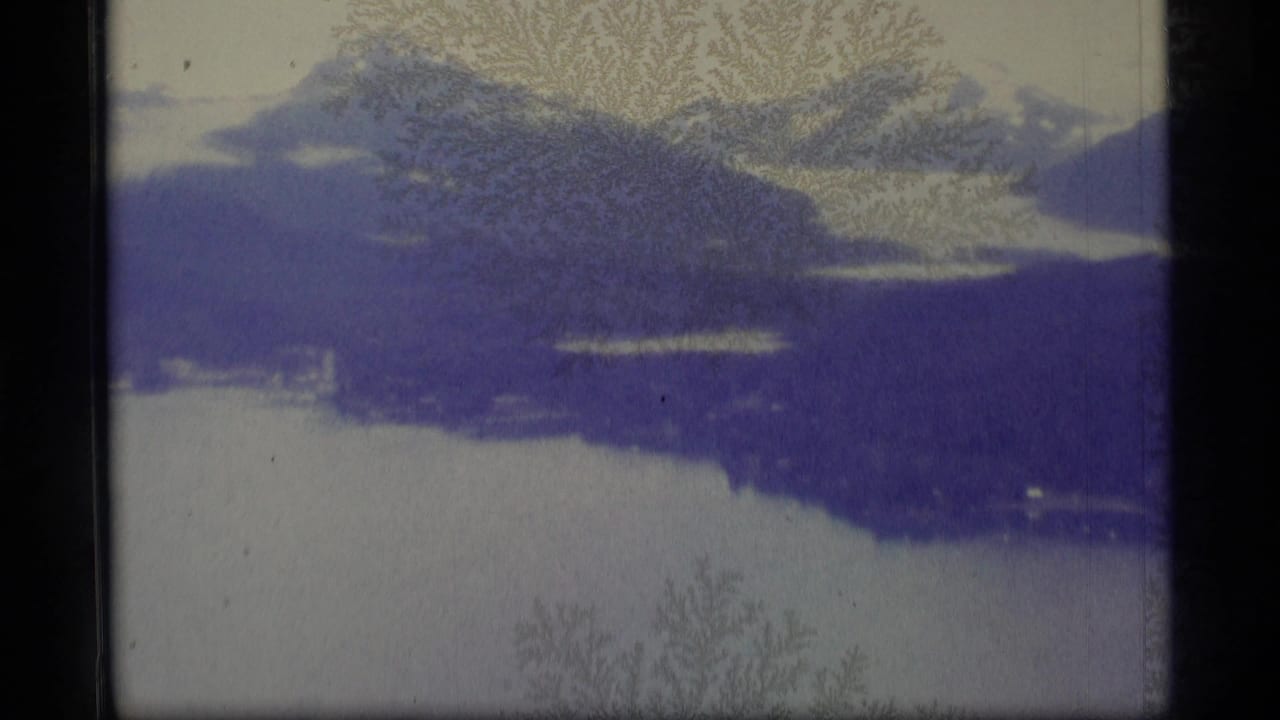 1977 Alaska home-movie frame showing snow-capped mountains above a glacial lake, softened by blue cast and frost-like film patterns.