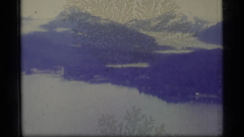 1977 Alaska home-movie frame showing snow-capped mountains above a glacial lake, softened by blue cast and frost-like film patterns.