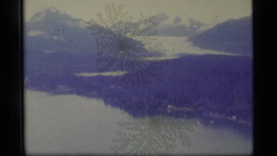 1977 Alaska 8mm frame of a mountain lake view with distant snowy peaks, dark forested slopes, and frost-like marks across the image.