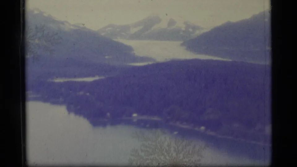 1977 Alaska home-movie frame showing a pale lake framed by dark slopes and distant snow-covered mountains, with soft 8mm grain.