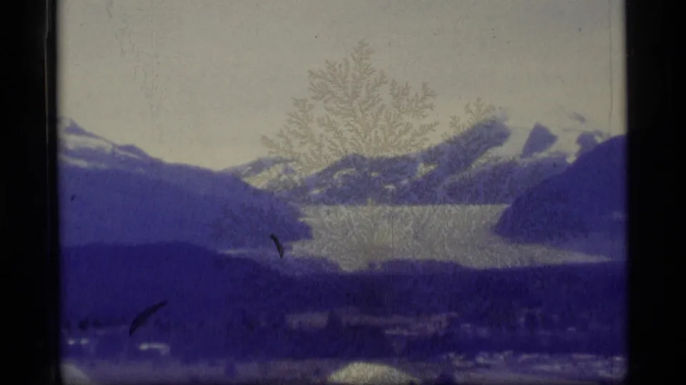 1977 Alaska 8mm frame showing a snow-capped ridge over a glacial lake, with dark foreground hills, visible scratches, and blue color shift.
