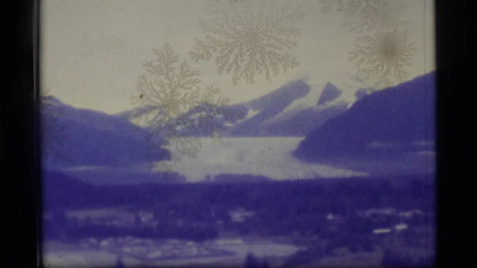 1977 Alaska home-movie frame with a wide lake, surrounding mountain slopes, snow patches, and soft 8mm film texture.