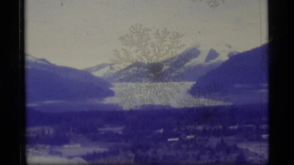 1977 Alaska 8mm frame showing a lake below snow-marked mountains, with dark wooded shoreline and visible film surface marks.