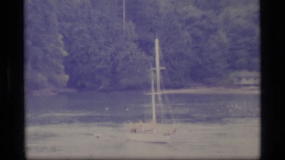1977 Alaska 8mm frame showing a small sailboat on calm lake water with dense evergreen trees along the shore.