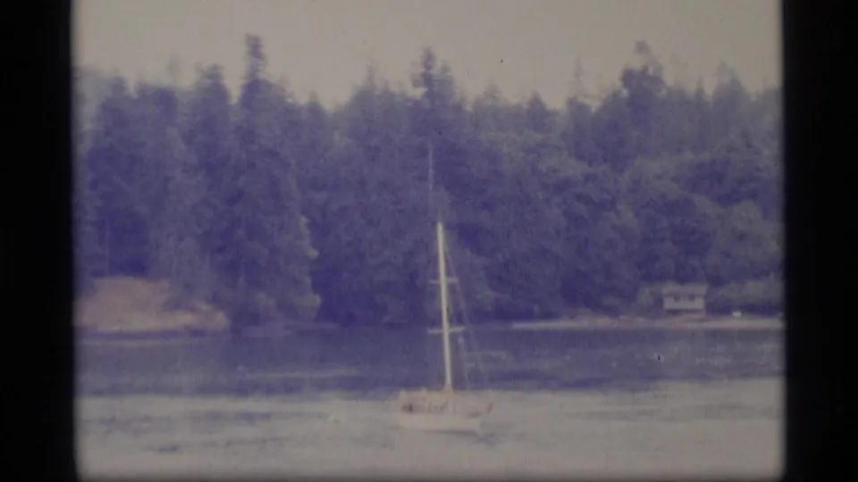 1977 Alaska 8mm frame showing a white sailboat in a sheltered bay below dense evergreen forest and a distant shoreline cabin.