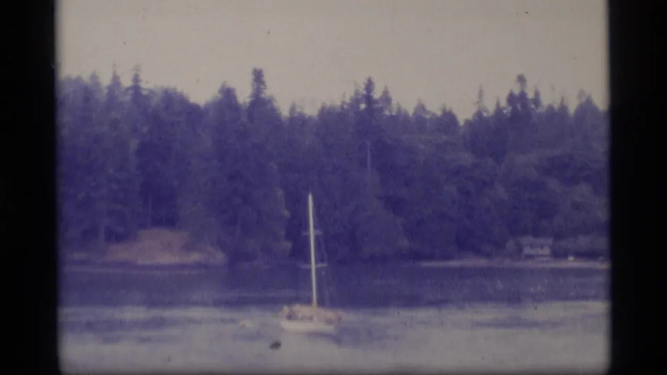 1977 Alaska home-movie frame with a sailboat crossing a quiet bay, backed by evergreen forest and a small distant shoreline structure.