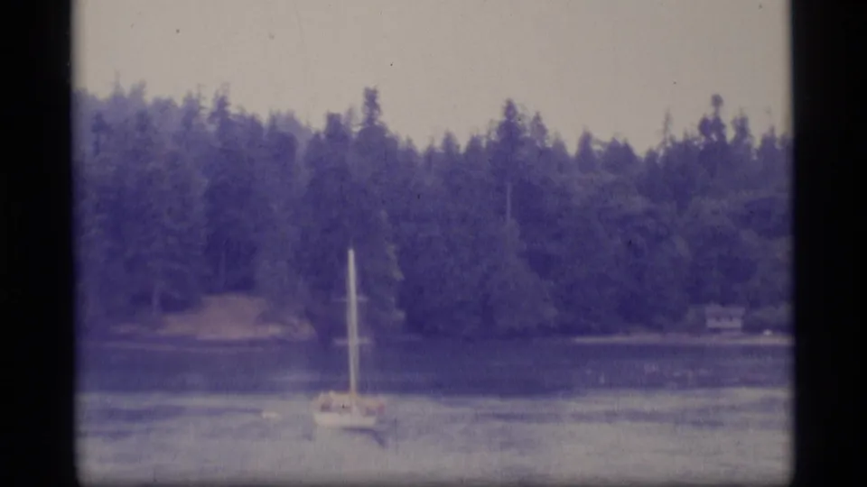 1977 Alaska 8mm frame showing a sailboat low in frame, calm water, dense conifers, and a small cabin along the far shore.