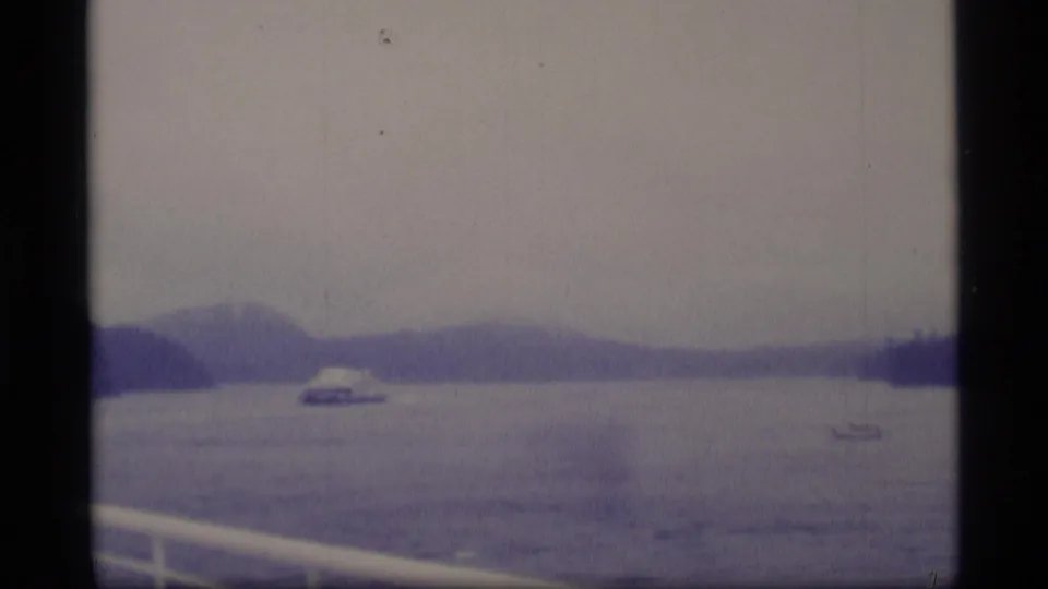Wide 1977 Alaska 8mm view from a moving vessel, with rail in foreground, a distant boat, calm water, and hazy low mountains.