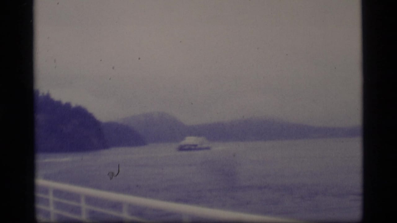 1977 Alaska 8mm frame showing a distant ferry crossing a broad gray waterway between forested mountain slopes, filmed from another boat.