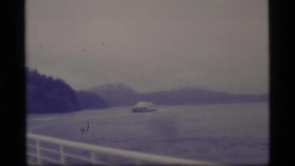 1977 Alaska 8mm frame showing a distant ferry crossing a broad gray waterway between forested mountain slopes, filmed from another boat.