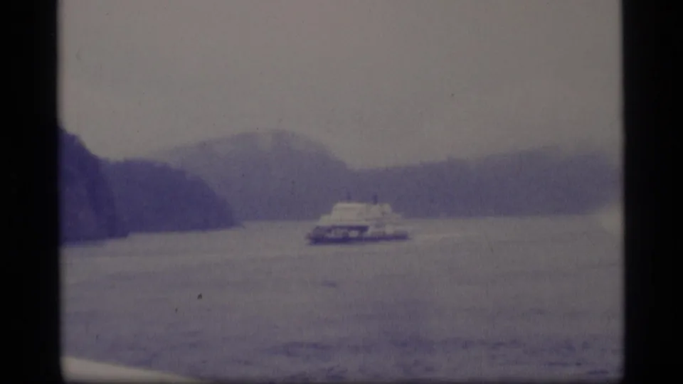 1977 Alaska home-movie frame with a white ferry moving across a misty mountain-lined waterway under an overcast sky.