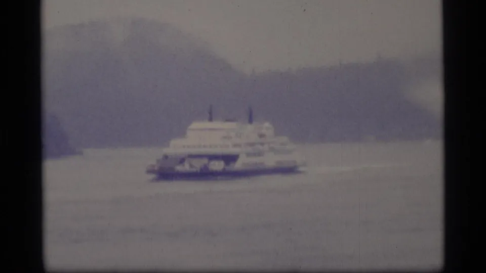 Soft 8mm view of a ferry passing across an Alaska waterway, with muted color and forested hills behind it.