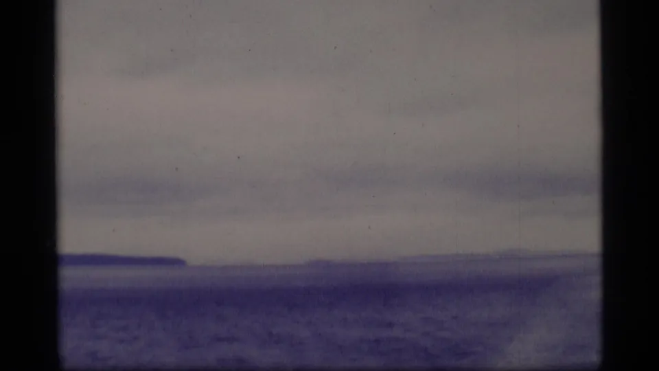 1977 Alaska 8mm view of a blue-gray lake or frozen surface under cloudy sky, with distant shoreline and dark frame edges.